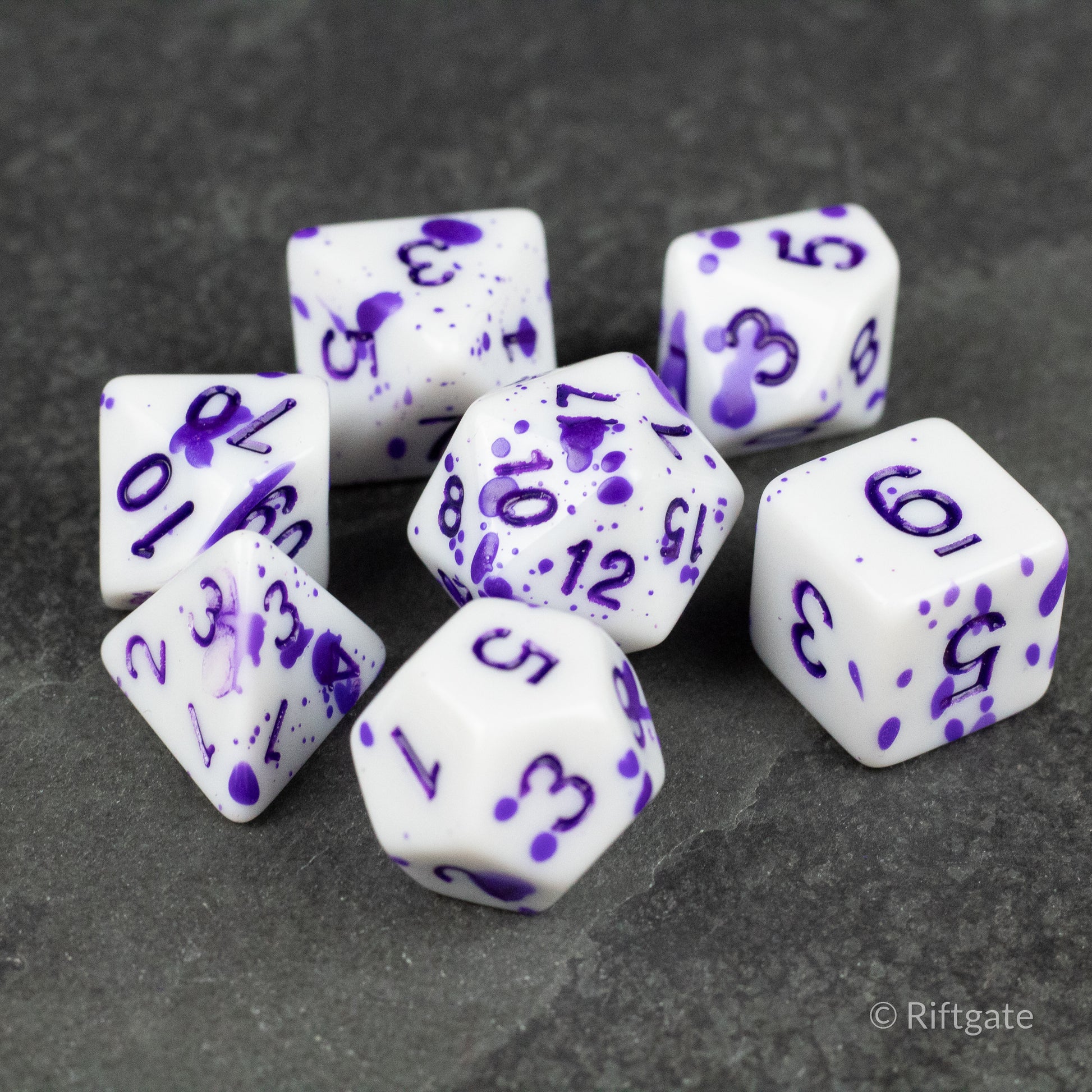 Color Change Purple Splatter Dice Set - Seven white polyhedral dice with purple splatter patterns and purple numbers on a dark background.