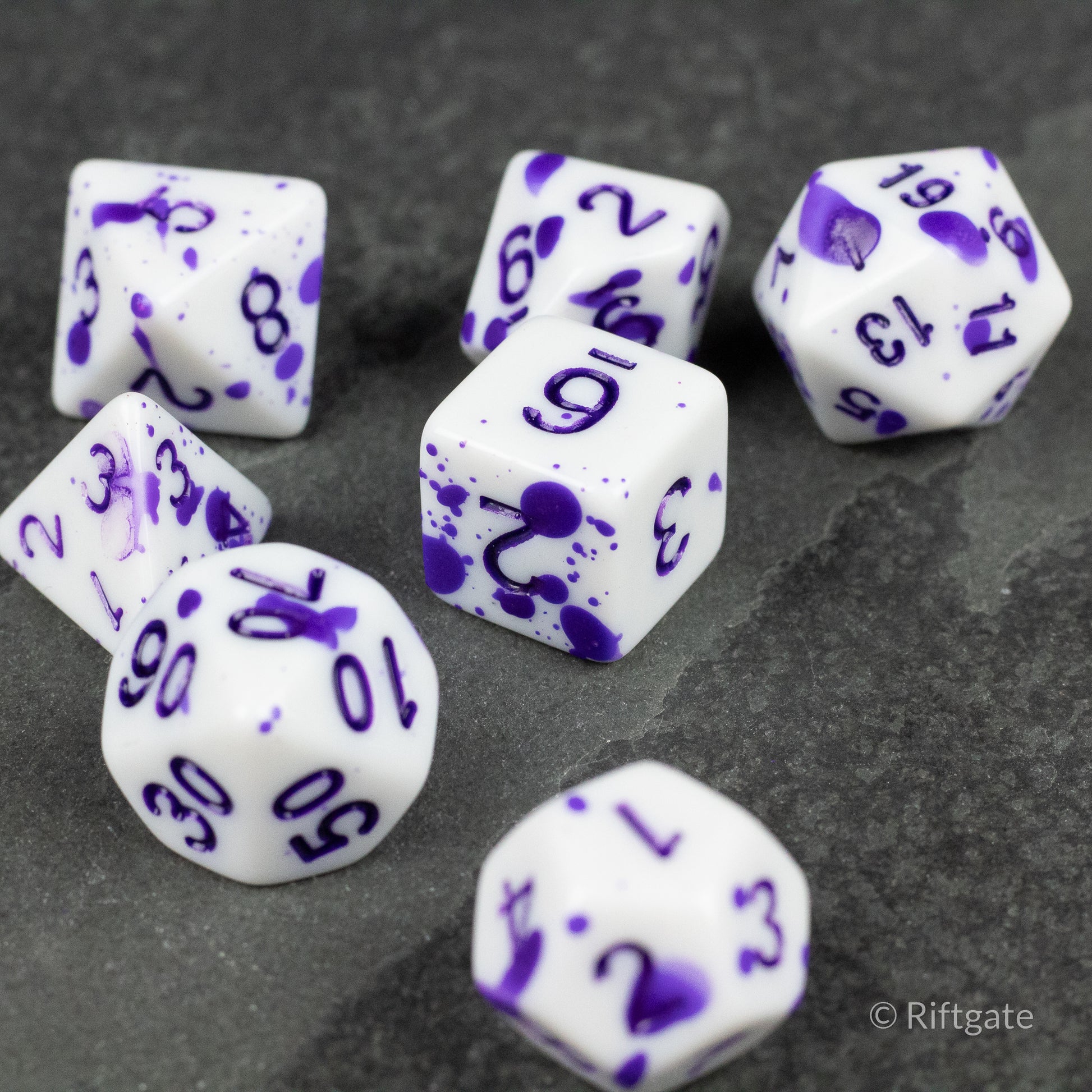 Color Change Purple Splatter Dice Set - Seven white polyhedral dice with purple splatter patterns and purple numbers, including d4, d6, d8, d10, d12, and d20 shapes.