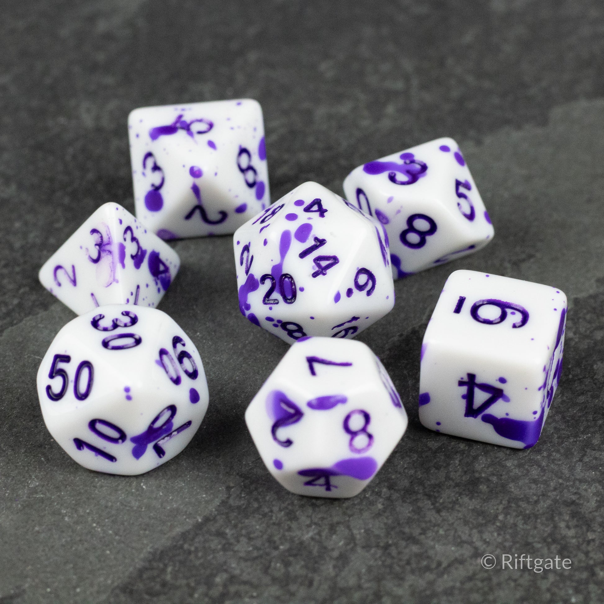 Color Change Purple Splatter Dice Set - Seven white polyhedral dice with purple splatter patterns and numbers, including d20, d12, d10, d8, d6, and d4 shapes.