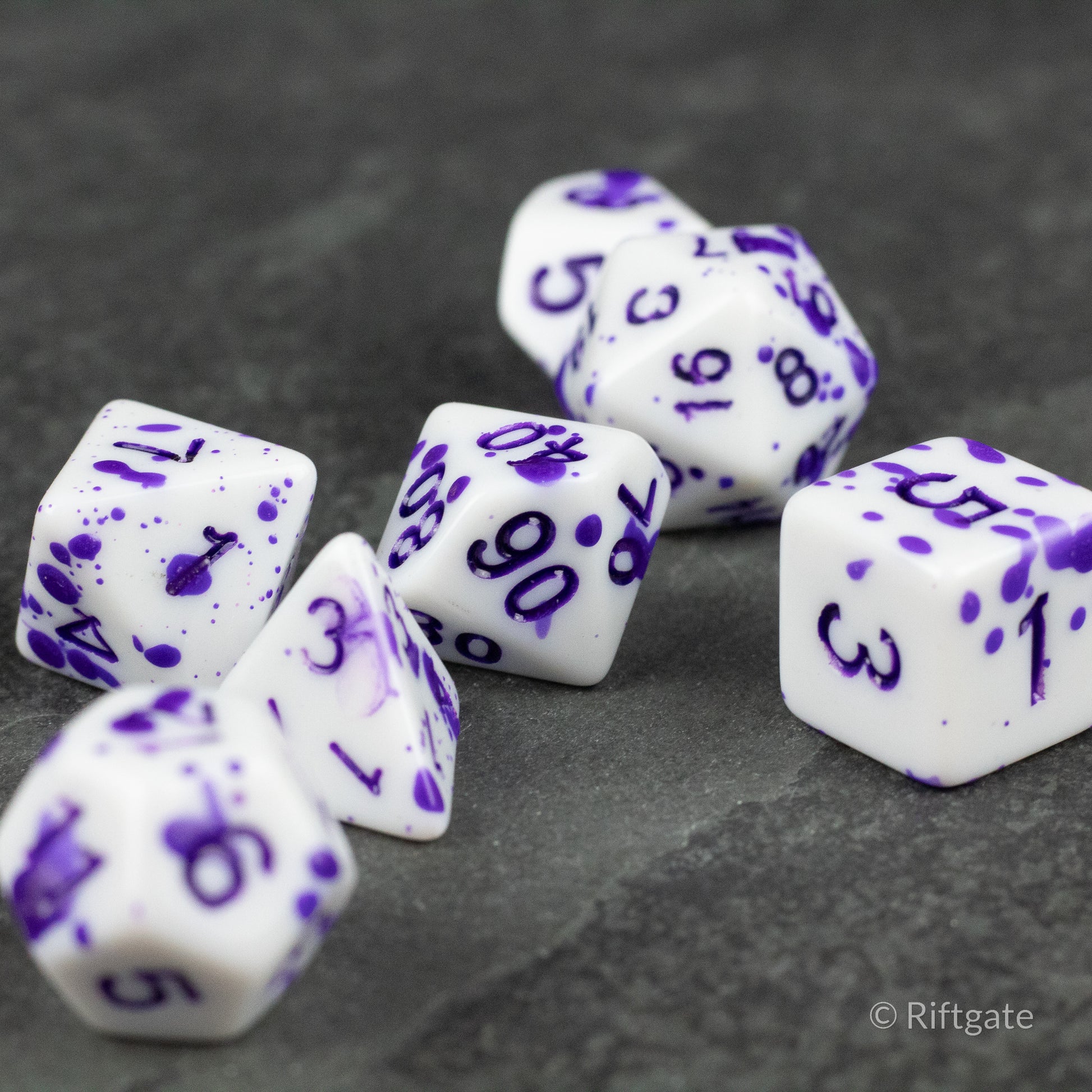 Color Change Purple Splatter Dice Set - White polyhedral dice with purple splatter patterns and purple numerals, set of seven on dark surface.