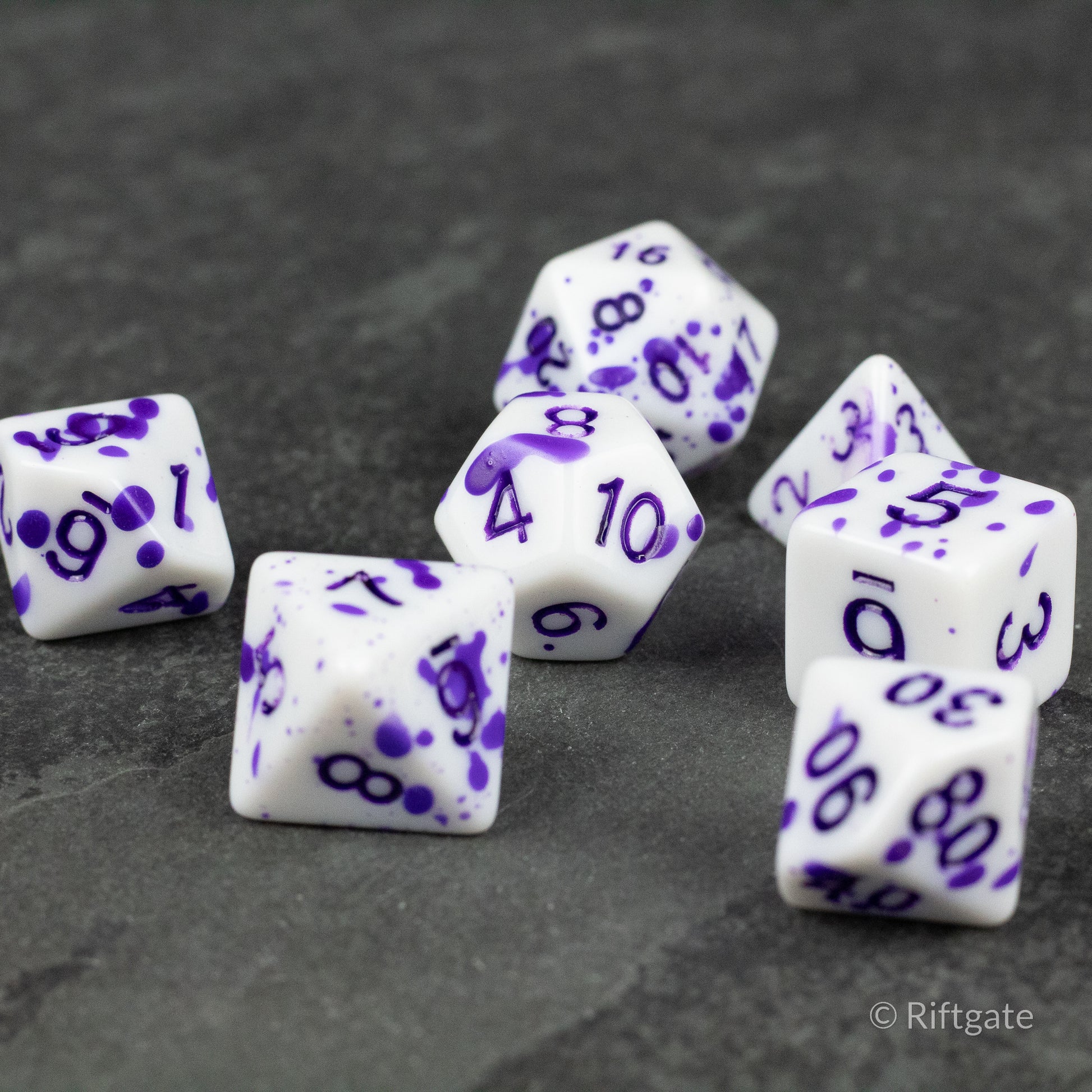 Color Change Purple Splatter Dice Set - Seven white polyhedral dice with purple ink splatter and numbers, featuring various shapes for tabletop gaming.