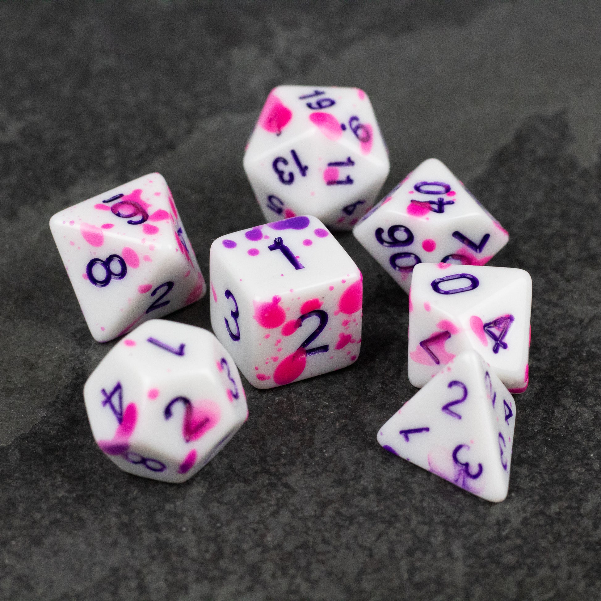 Color Change Purple Splatter Dice Set - Seven white polyhedral dice with purple numbers and pink and purple splatter patterns on a dark surface.