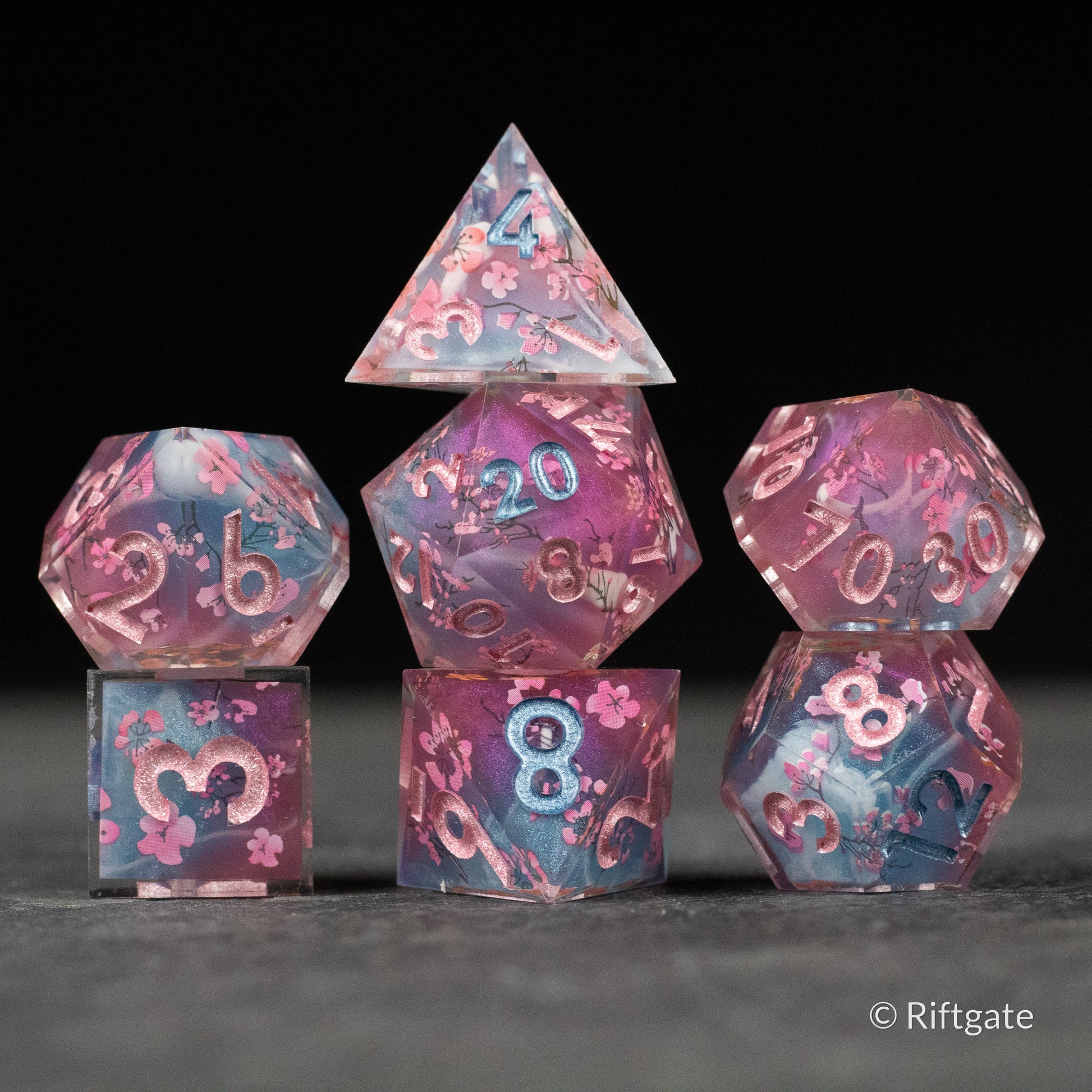 Twilight Blossoms Dice Set - Set of seven sharp-edged polyhedral dice with pink and blue marbled pattern and metallic rose gold numbers.