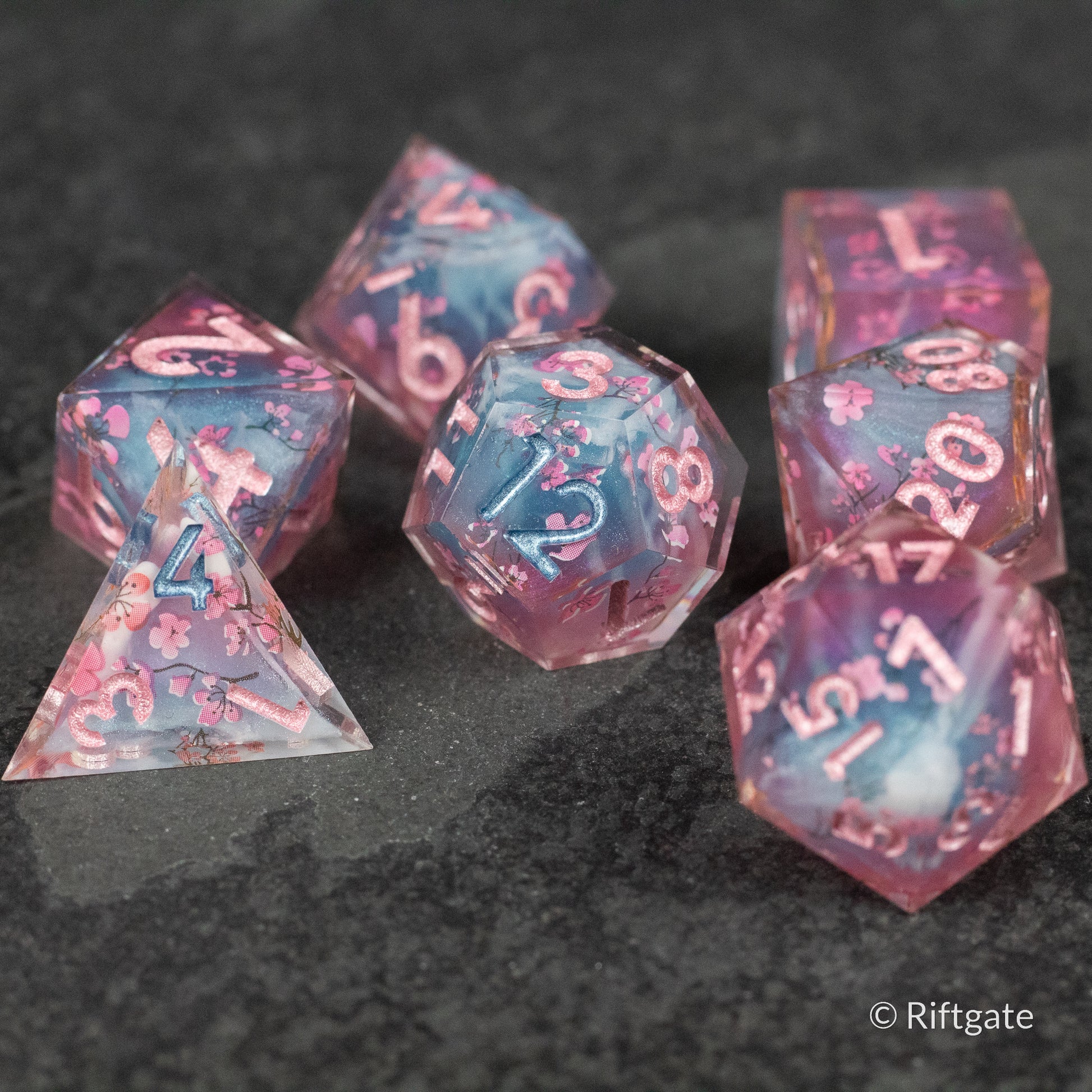 Twilight Blossoms Dice Set - Translucent pink and blue sharp-edged polyhedral dice set with white numbering on dark gray surface.