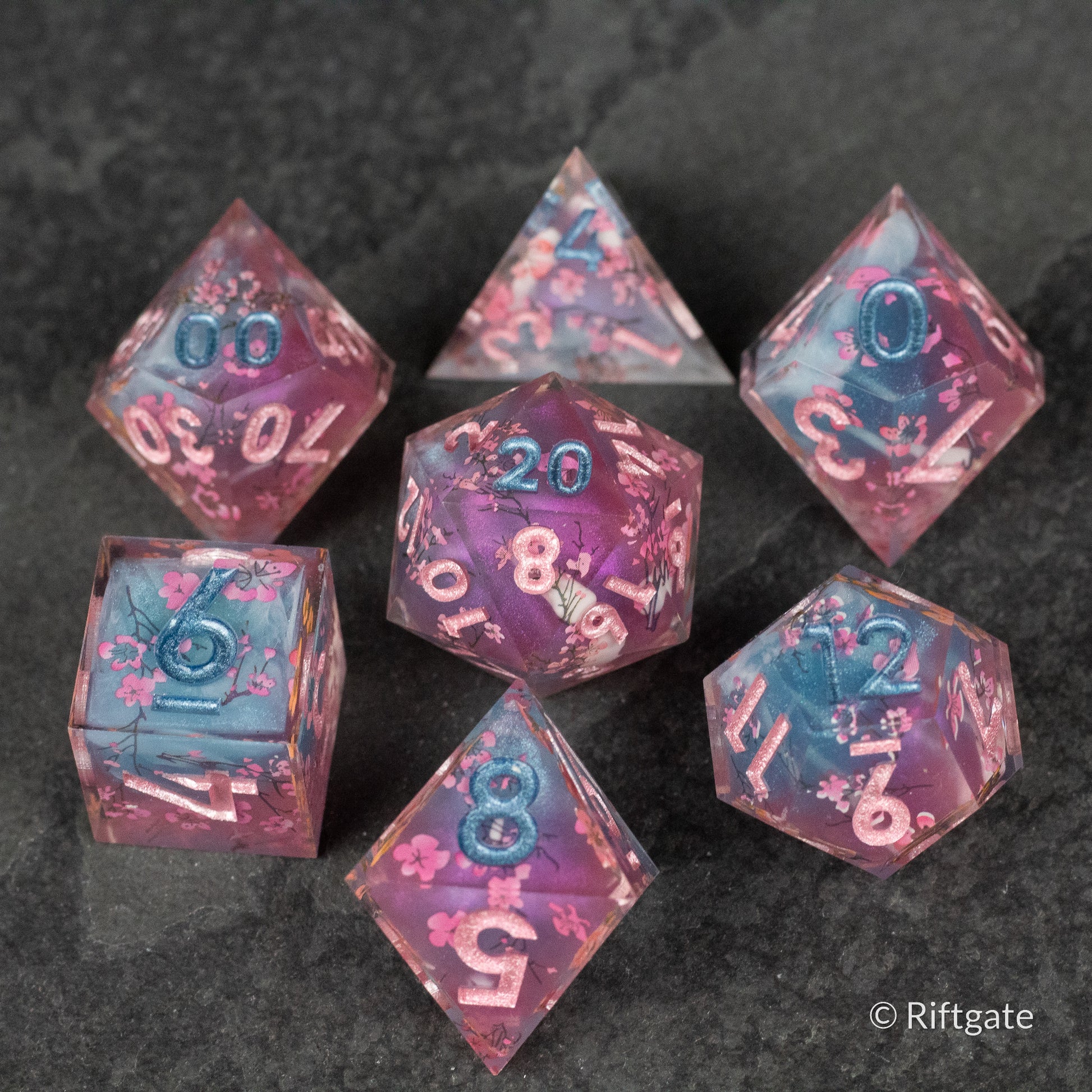 Twilight Blossoms Dice Set - Seven pink and blue translucent polyhedral dice with sharp edges and white numbering, featuring floral swirls inside.