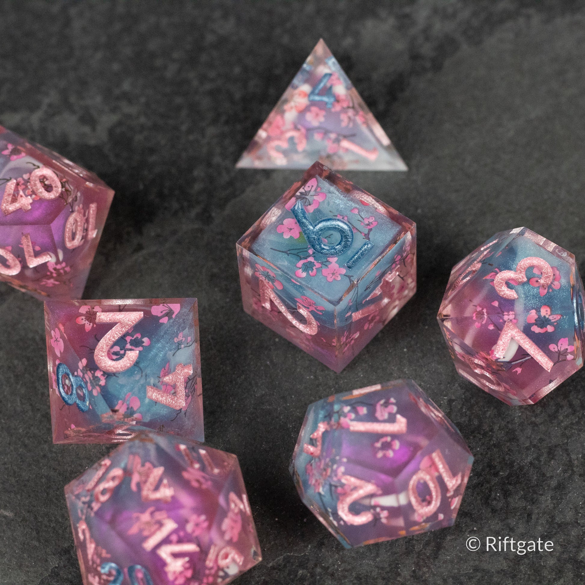 Twilight Blossoms Dice Set - Set of translucent purple and blue polyhedral dice with pink floral patterns and sharp edges, numbered in white.