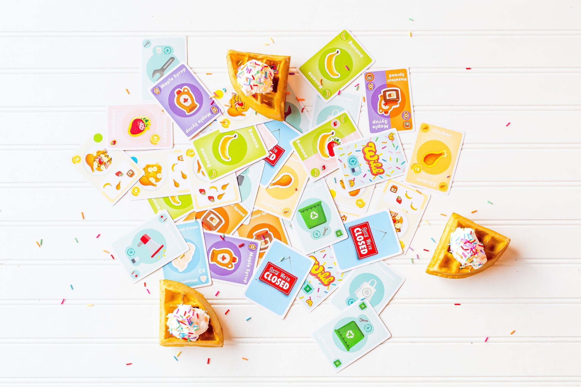 Top Waffle - Colorful playing cards scattered around small waffle cones topped with whipped cream and sprinkles on a white wooden surface.