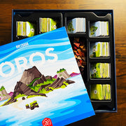 Oros Collector's Edition - Stacked hexagonal game tiles with colorful landscapes organized in black trays next to a blue strategy game box featuring.