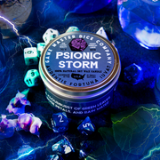 Psionic Storm Gaming Candle - Round blue candle tin with purple dice logo labeled Psionic Storm surrounded by colorful gaming dice on a dark.