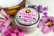 Stillness of Mind Gaming Candle - Round pink metal tin candle with "Stillness of Mind" label surrounded by pink dice and flowers on white surface.
