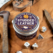 Studded Leather Gaming Candle - Round brown tin candle with a studded leather label, surrounded by metal and plastic polyhedral dice on a tan leather.