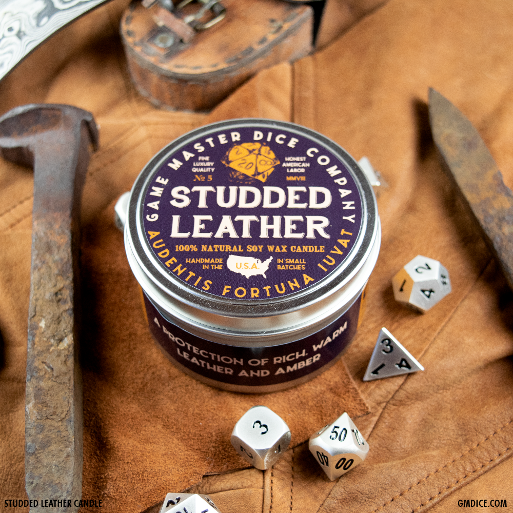 Studded Leather Gaming Candle - Round brown tin candle with a studded leather label, surrounded by metal and plastic polyhedral dice on a tan leather.