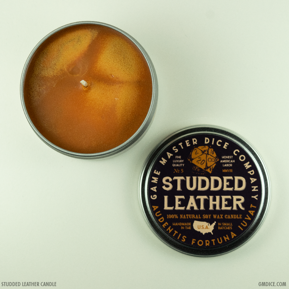 Studded Leather Gaming Candle - Round orange-brown candle in a silver tin with black lid labeled Studded Leather by Game Master Dice Company.