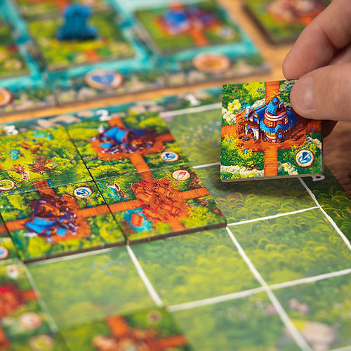 Timber Town - Colorful board game with forest and village tiles, featuring a hand placing a small detailed tile with a blue building and.