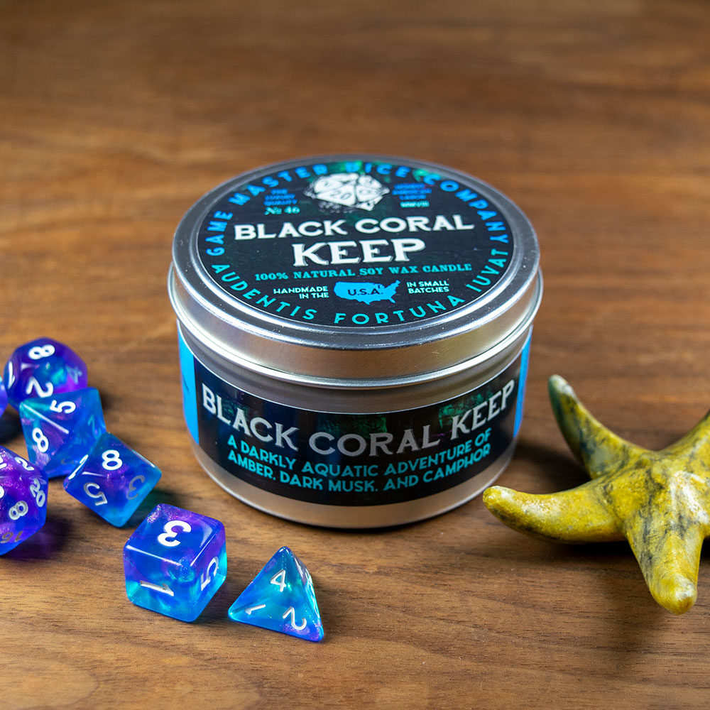 Black Coral Keep Candle - Small round metal tin candle with black and turquoise label reading Black Coral Keep, placed next to colorful dice and a.