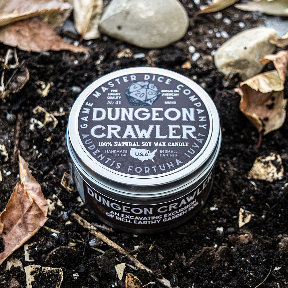 Dungeon Crawler Gaming Candle - Black cylindrical gaming candle with white bold text reading "Dungeon Crawler," natural soy wax, placed on soil with leaves.