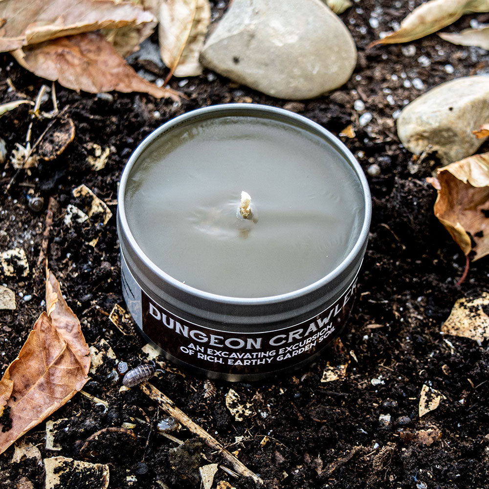 Dungeon Crawler Gaming Candle - Black tin candle labeled Dungeon Crawler with white text, sitting on dark soil surrounded by dry leaves and stones.