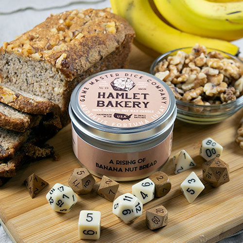 Hamlet Bakery Gaming Candle - Small round candle tin with pink label reading Hamlet Bakery, surrounded by banana slices, nuts, wooden dice, and rustic.