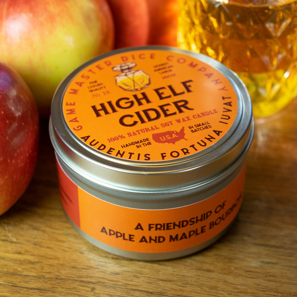 High Elf Cider Gaming Candle - Orange metal tin candle with a silver lid, labeled "High Elf Cider," featuring apple and maple scents on a wooden surface.