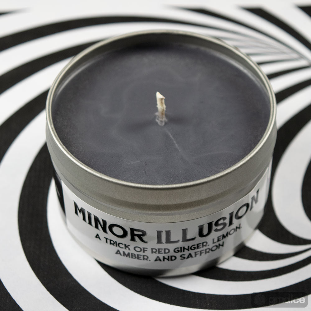 Minor Illusion Gaming Candle - Small round tin candle with black wax, labeled "Minor Illusion" with scent notes of red ginger, lemon, amber, and saffron,.