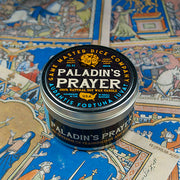 Paladin's Prayer Gaming Candle - Black round tin candle labeled Paladin's Prayer with gold and white text, resting on colorful medieval-themed gaming map.