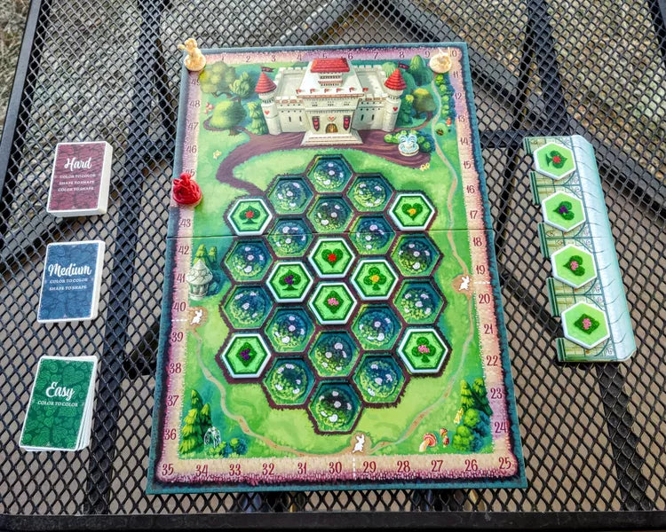 Paint the Roses: Deluxe Edition - Colorful board game setup with hexagonal green and blue tiles, castle illustration, cards, and tokens on mesh table.