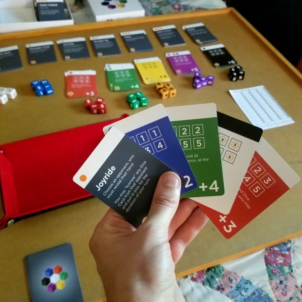 Bare Bones - Hand holding five colorful game cards with numbers and symbols, set against a wooden game board with matching multicolored.