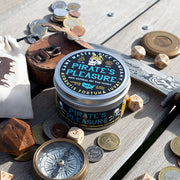 Pirate's Pleasure Gaming Candle - Round black metal tin with teal and gold Pirate's Pleasure label, surrounded by coins, dice, and wooden gaming accessories.