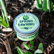 Spring Dawning Gaming Candle - Round green tin candle with "Spring Dawning" label nestled among green plant leaves and mulch.
