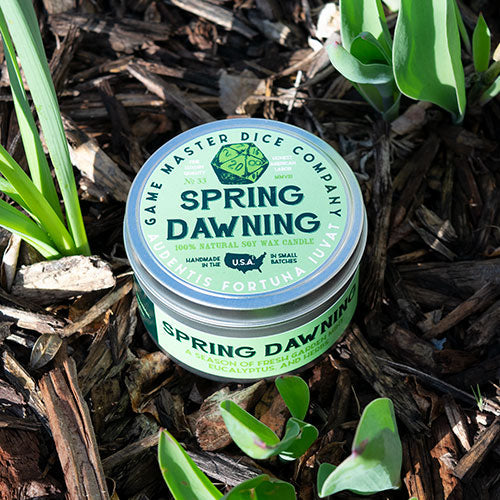 Spring Dawning Gaming Candle - Round green tin candle with "Spring Dawning" label nestled among green plant leaves and mulch.
