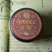 Hobbit Leaf Gaming Candle - Round dark purple tin candle with gold lettering and a d20 die graphic on the lid labeled "Hobbit Leaf".
