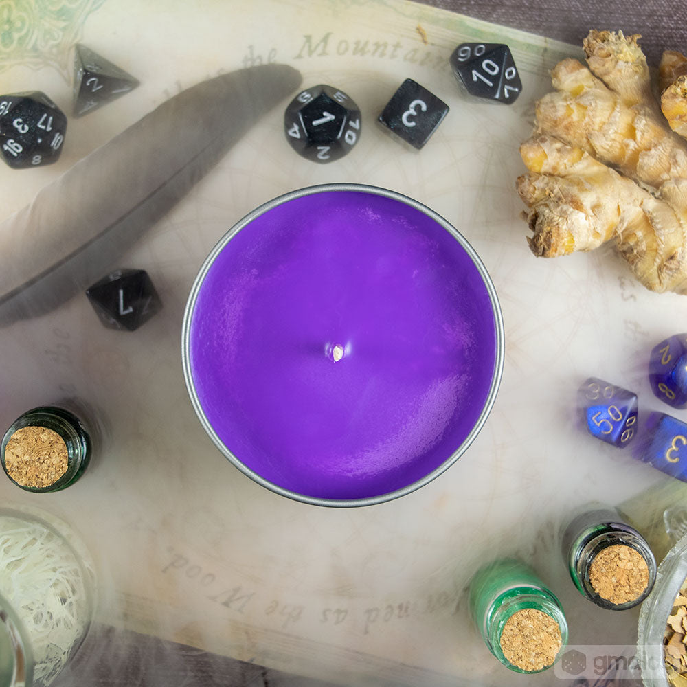 Toil and Trouble Candle - Round purple candle in metal tin surrounded by black and blue dice, ginger root, and small glass bottles with cork stoppers.