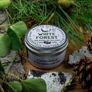 White Forest Gaming Candle - Round metal tin candle with white label featuring green forest illustration and text, placed on natural wood with green.