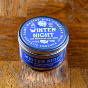 Winter Night Gaming Candle - Small round tin candle with blue label featuring white text and a winter-themed emblem, set on a wooden surface.