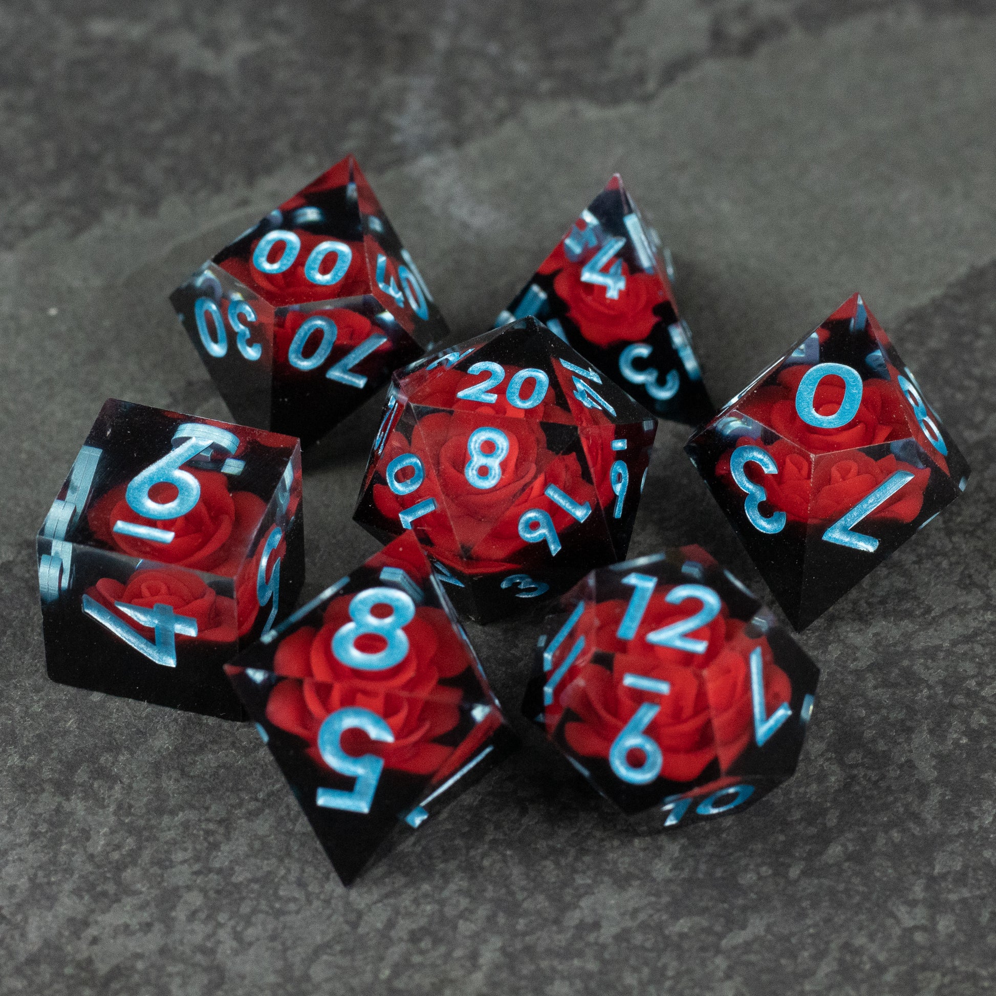 Cursed Rose Dice Set - Seven red and black marbled sharp-edged dice with light blue numbering on a gray textured surface.