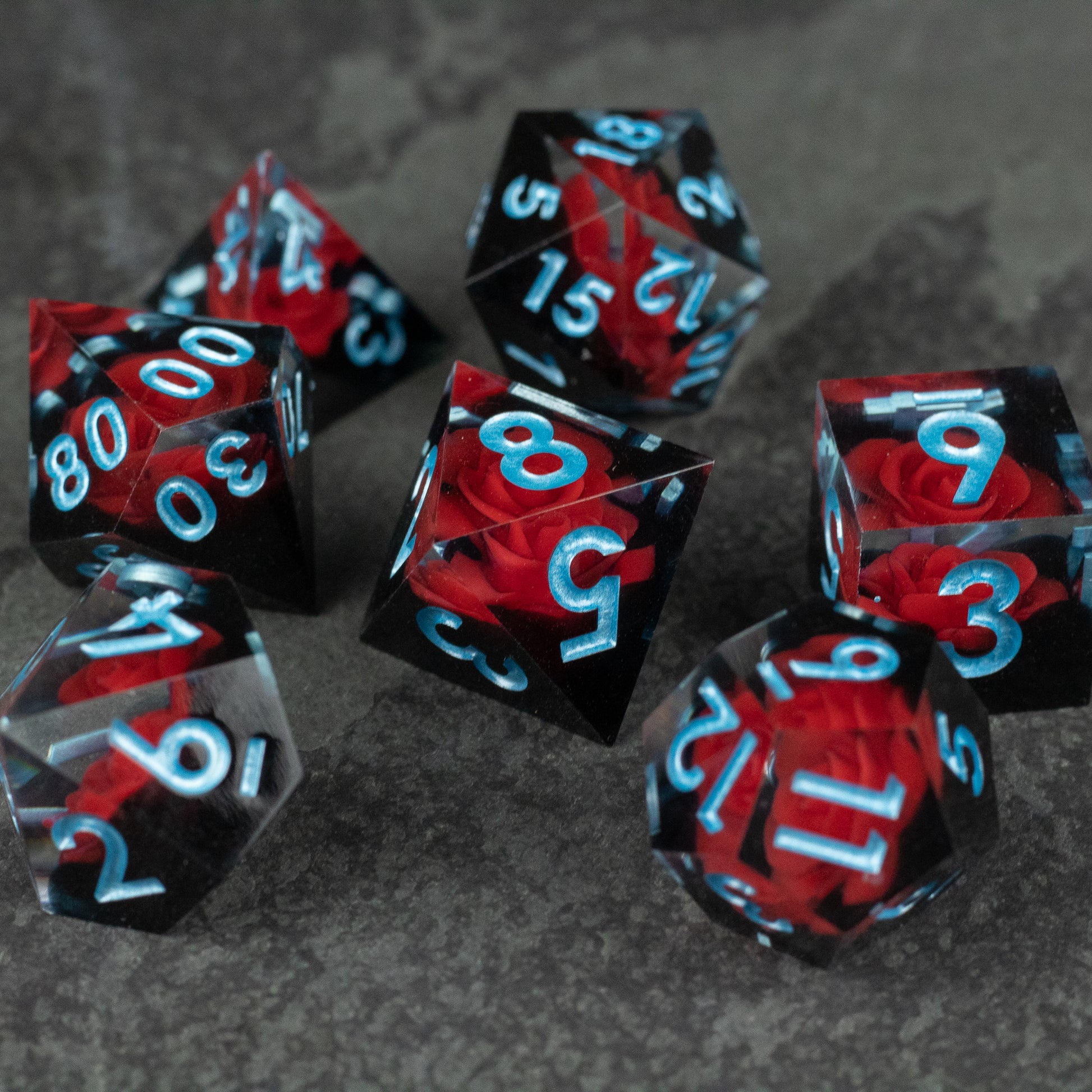 Cursed Rose Dice Set - Seven sharp-edged polyhedral dice in translucent red and black with sky blue numbers.