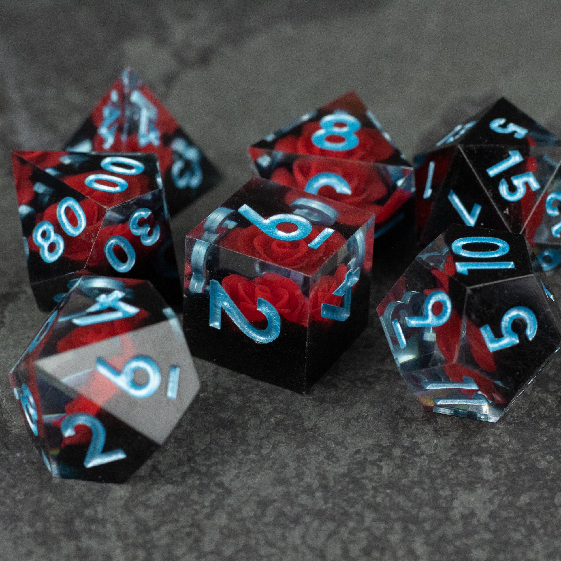 Cursed Rose Dice Set - Set of seven sharp-edged polyhedral dice with red rose designs inside, black and clear casing, and light blue numbers.