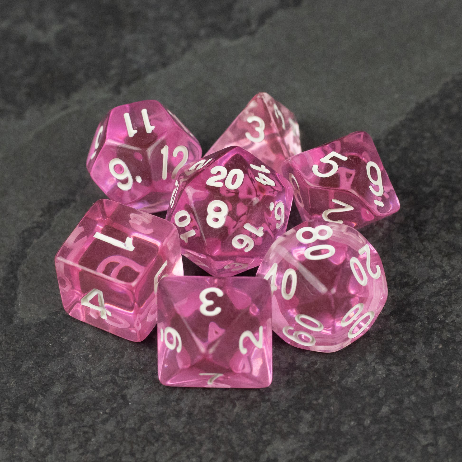 Pink Transparent Dice Set - Set of seven pink transparent polyhedral dice with white numbers on a dark textured surface.