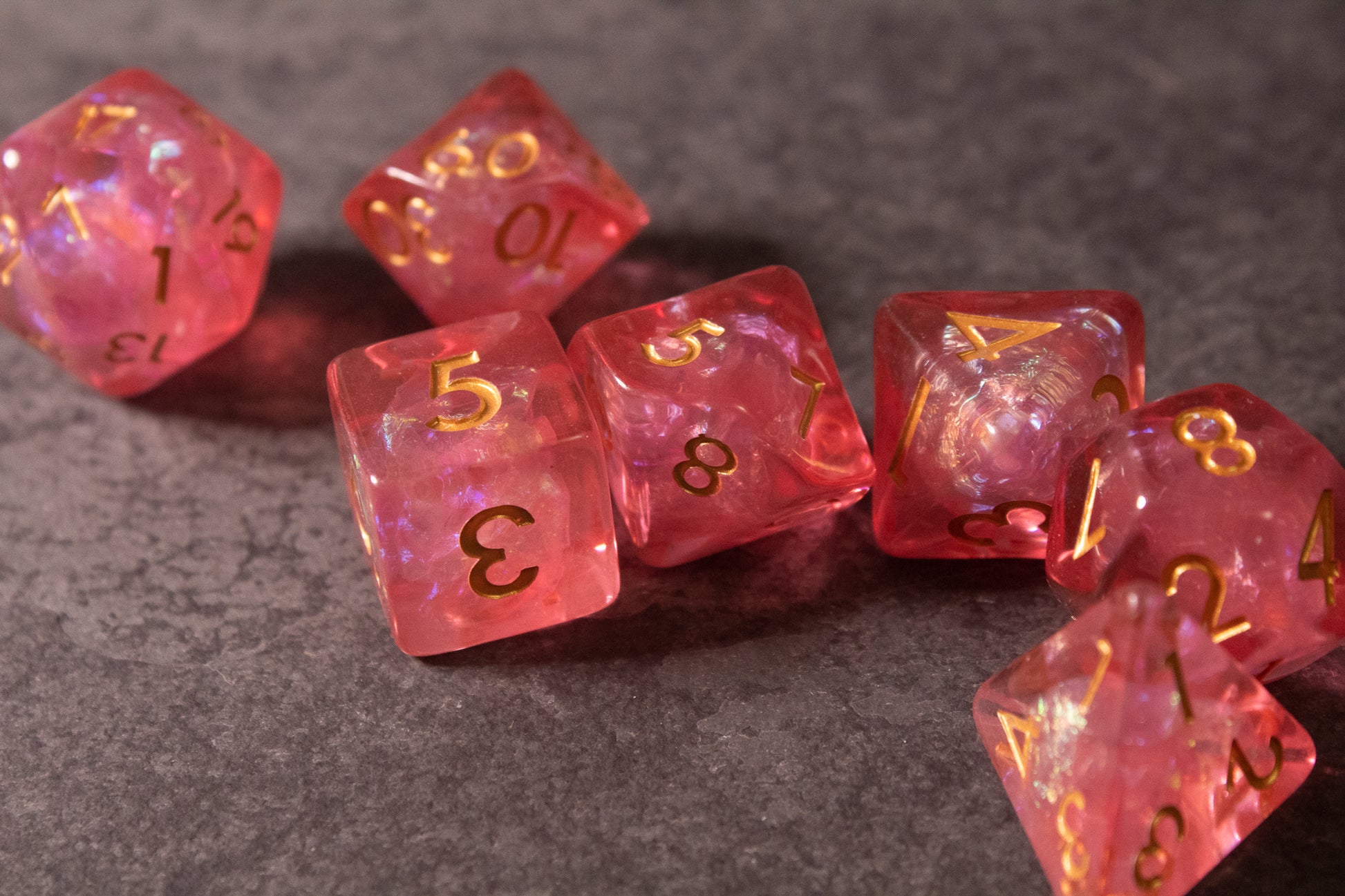 Pink Iridescent Dice Set - Pink iridescent seven-piece dice set with gold-toned numbers on gray surface.