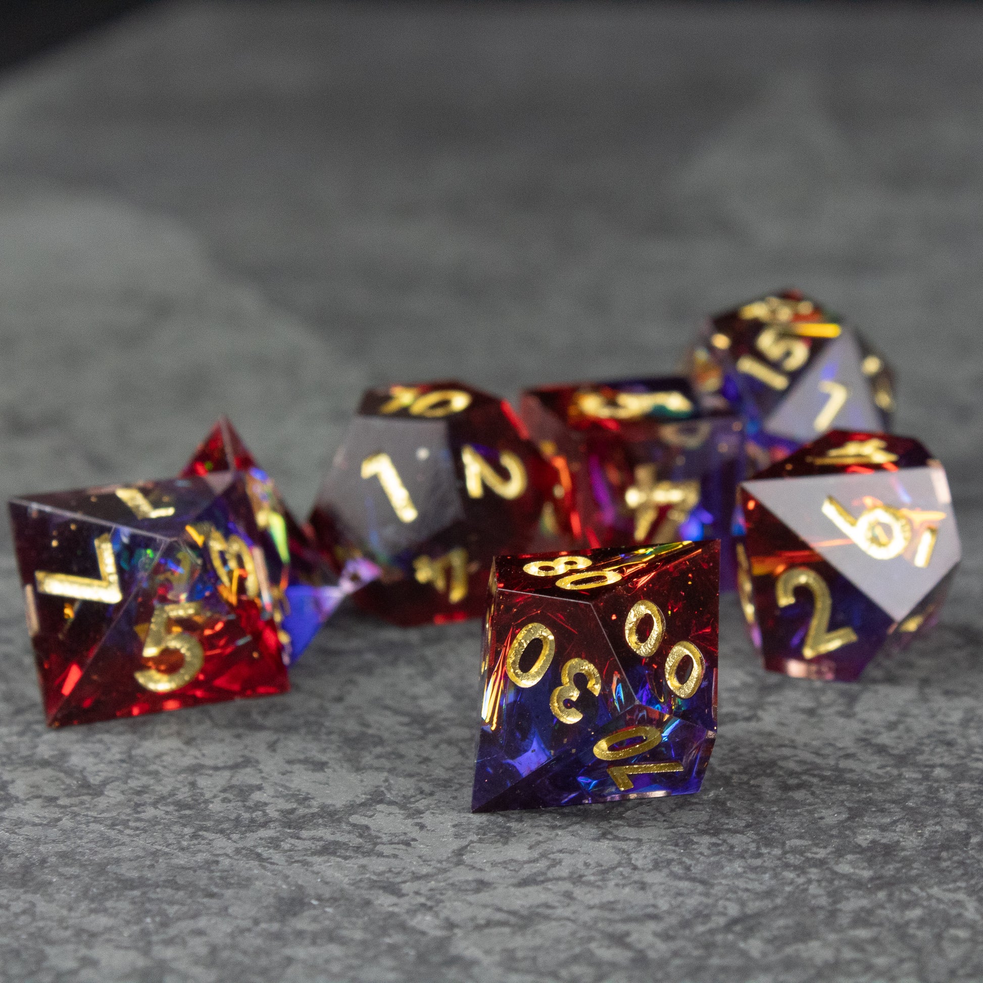 Electromancer Dice Set - Set of transparent blue and red sharp-edged polyhedral dice with gold numbers on gray surface.