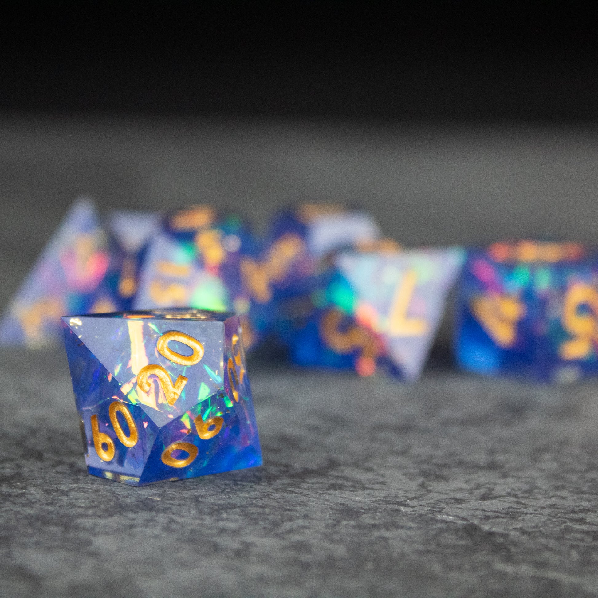 Arcana Dice Set - Blue translucent sharp-edged polyhedral dice with gold numbering on gray surface.