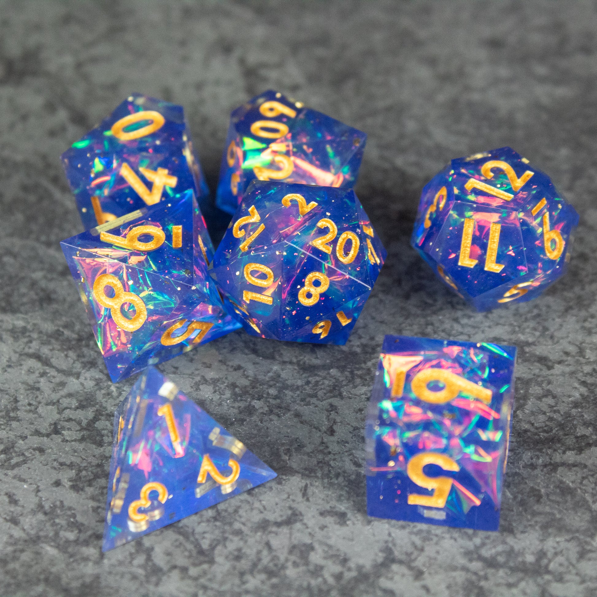 Arcana Dice Set - Set of translucent blue polyhedral dice with sharp edges and gold numbering, embedded with colorful glitter flecks.