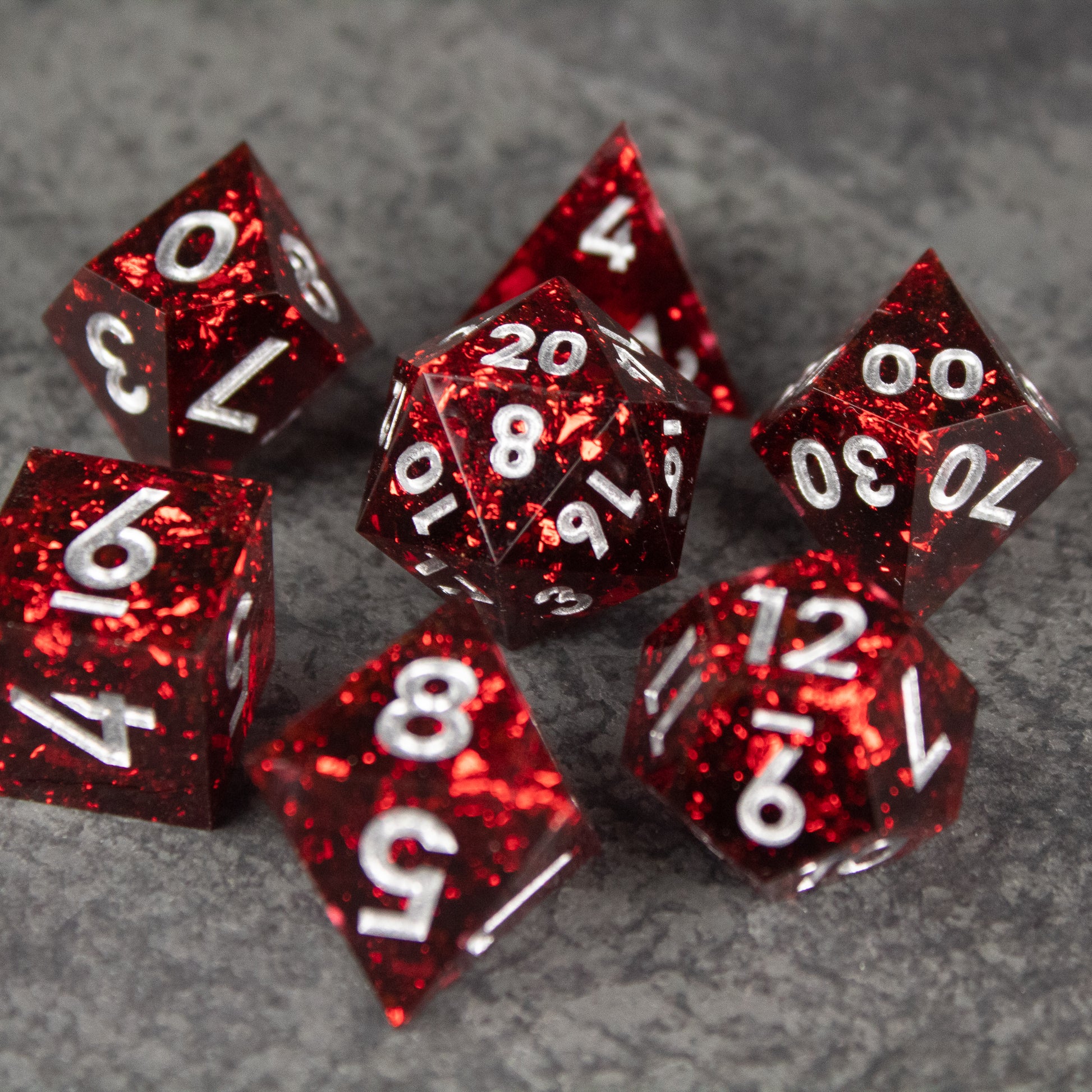 Inferno Dice Set - Set of seven sharp-edged translucent red dice with white numbers, including d20, d12, d10, d8, d6, and d4 on gray surface.