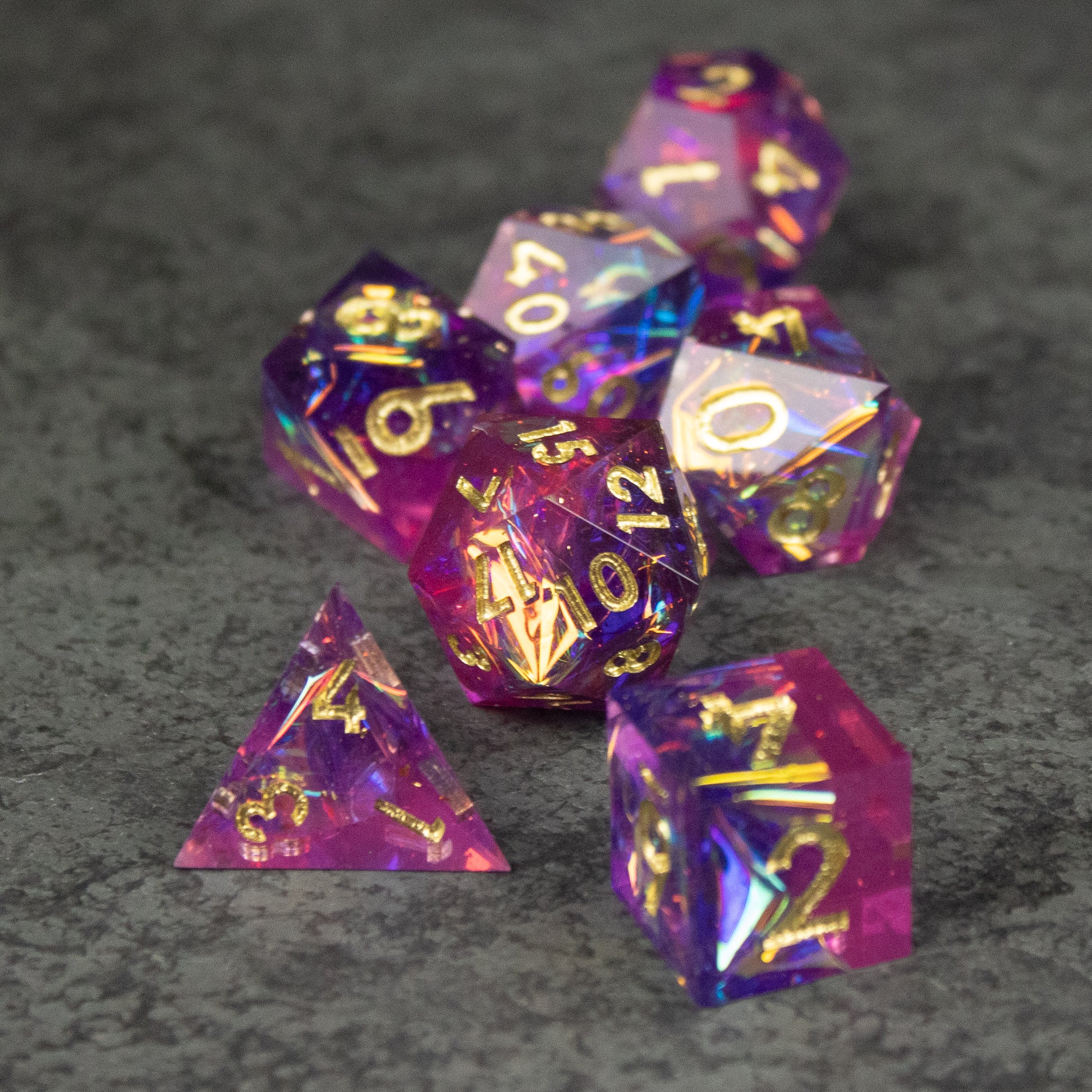 Aether Dice Set - Seven polyhedral dice in translucent pink and purple with sharp edges and gold numbering on a gray textured surface.