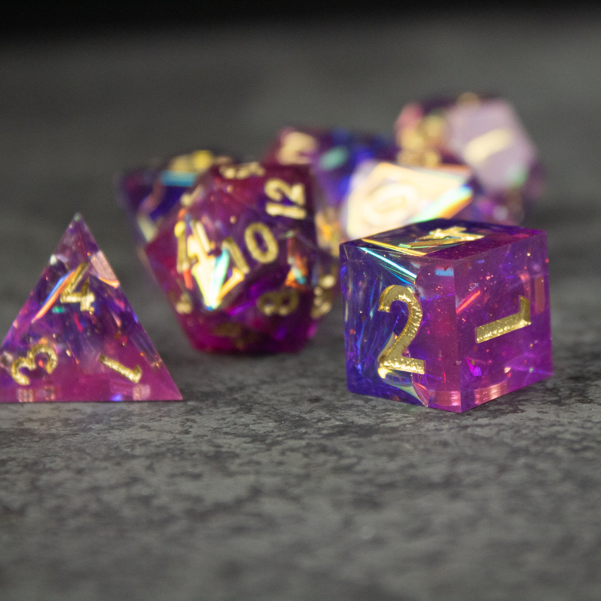Aether Dice Set - Purple and pink translucent sharp-edged polyhedral dice with gold numbers on gray surface.