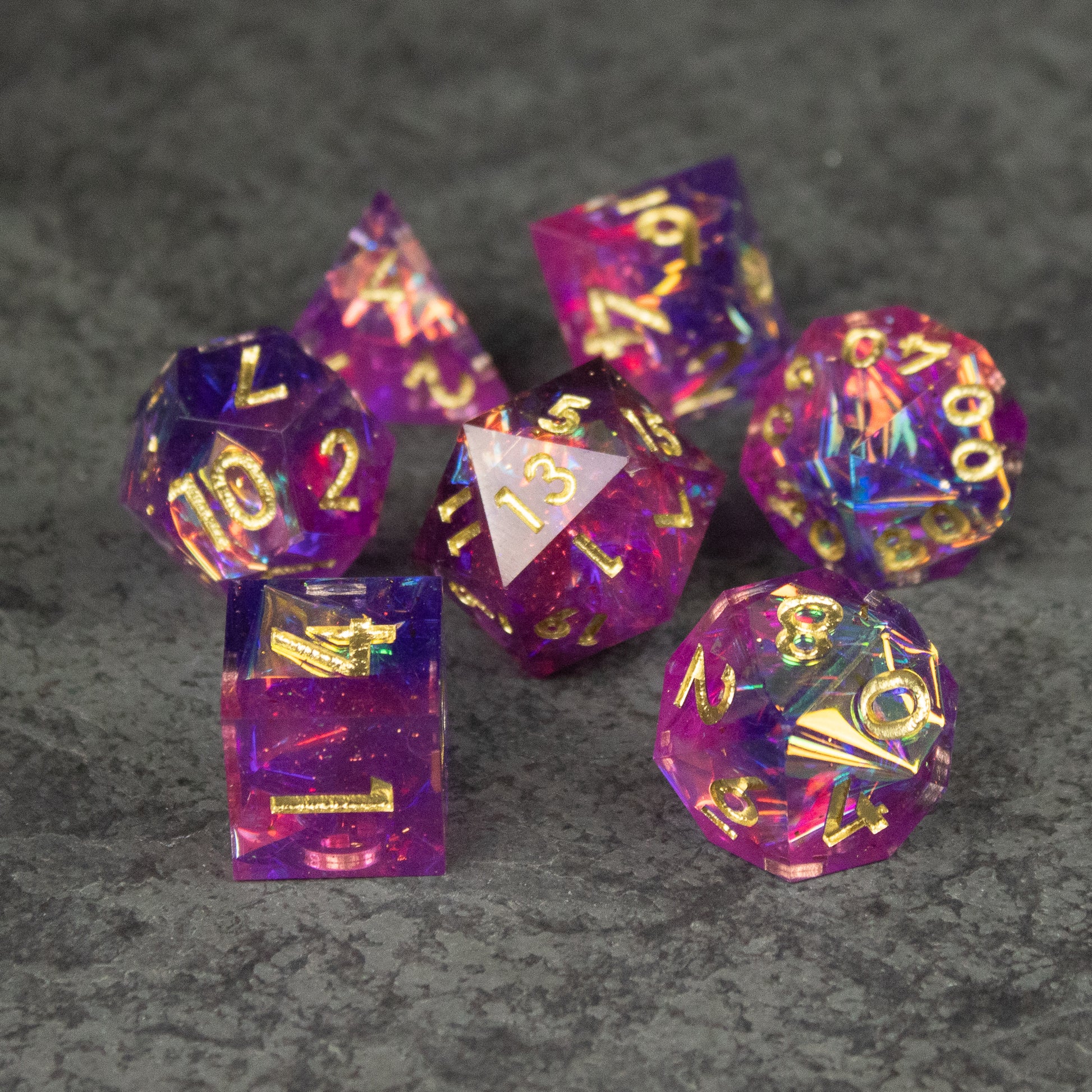 Aether Dice Set - Seven sharp-edged polyhedral dice in translucent pink and purple with gold numbering on a gray surface.