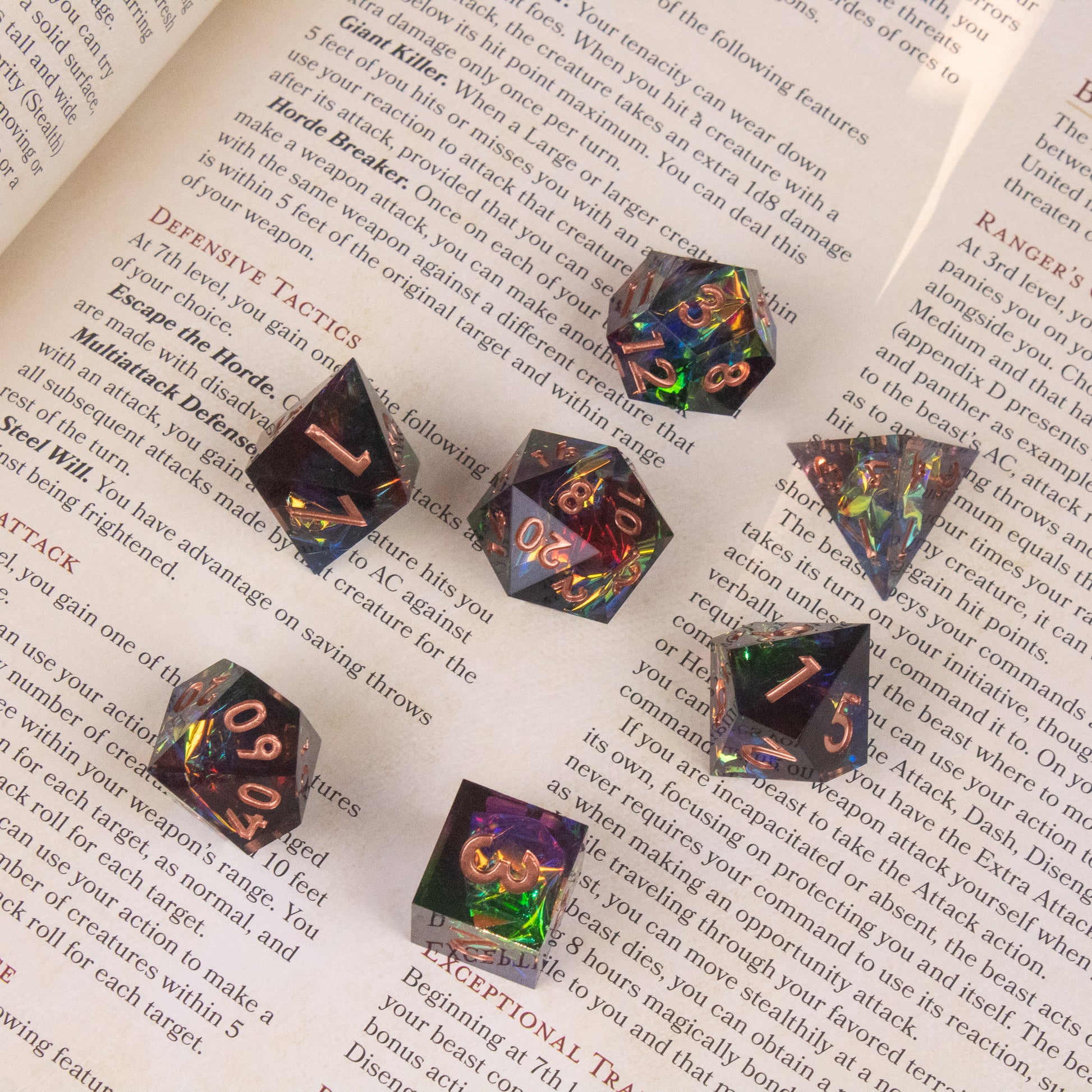 Aurora Dice Set - Seven sharp-edged polyhedral dice with a rainbow iridescent finish and gold numbers on a book page background.