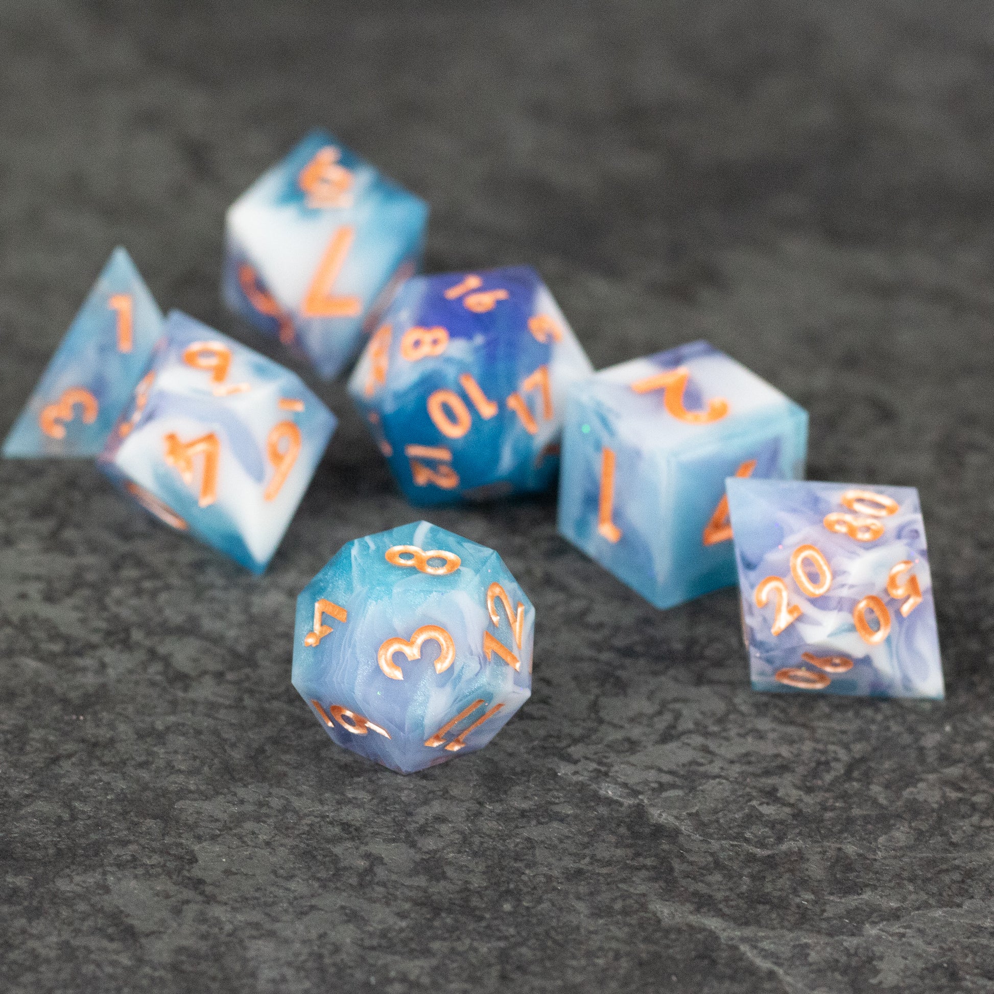 Cloudscape Dice Set - Set of seven sharp-edged dice in blue and white marbled pattern with orange numbers on a dark textured surface.