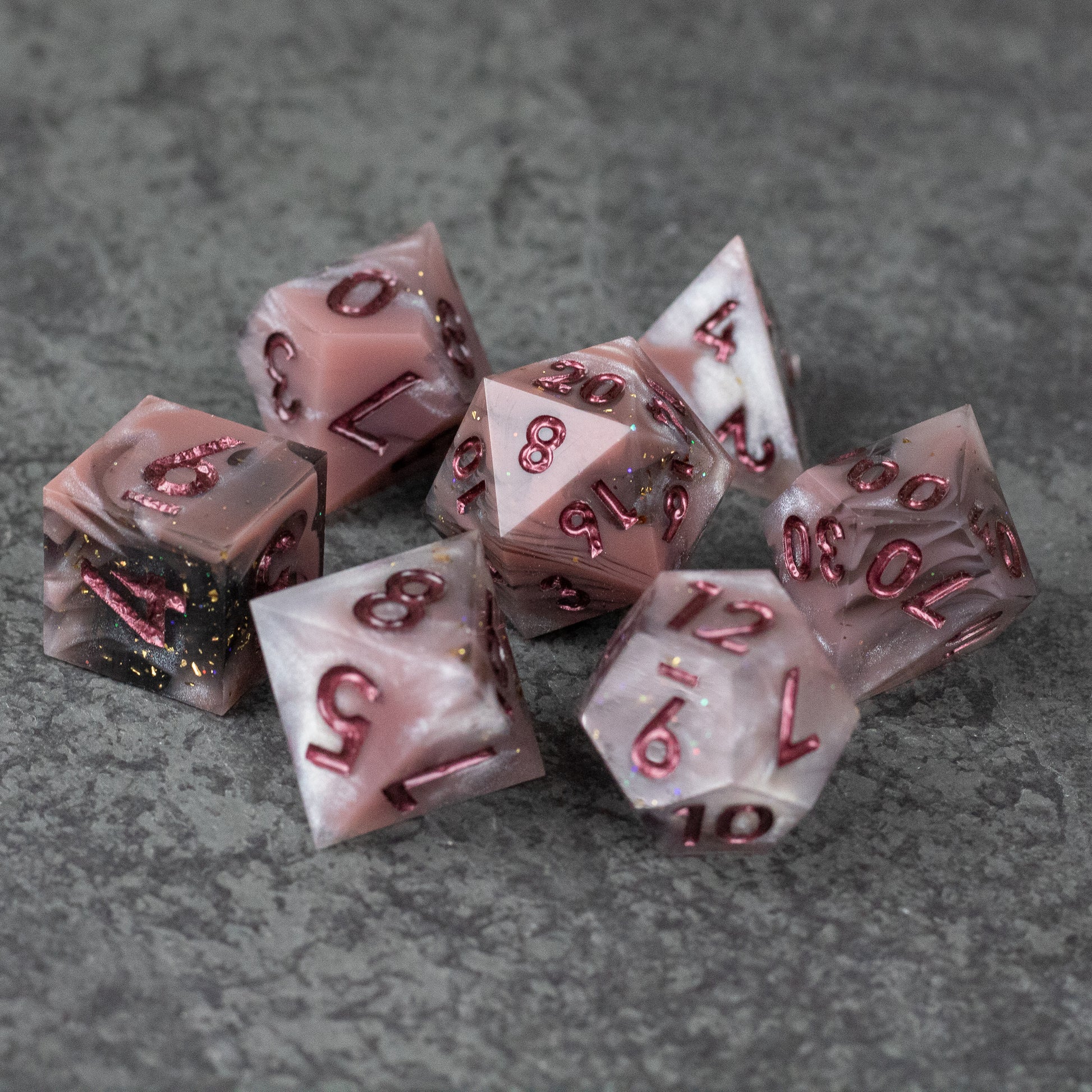 Rosestone Dice Set - Set of seven red and white marbled sharp edge dice with gold and red numbering on gray textured surface.