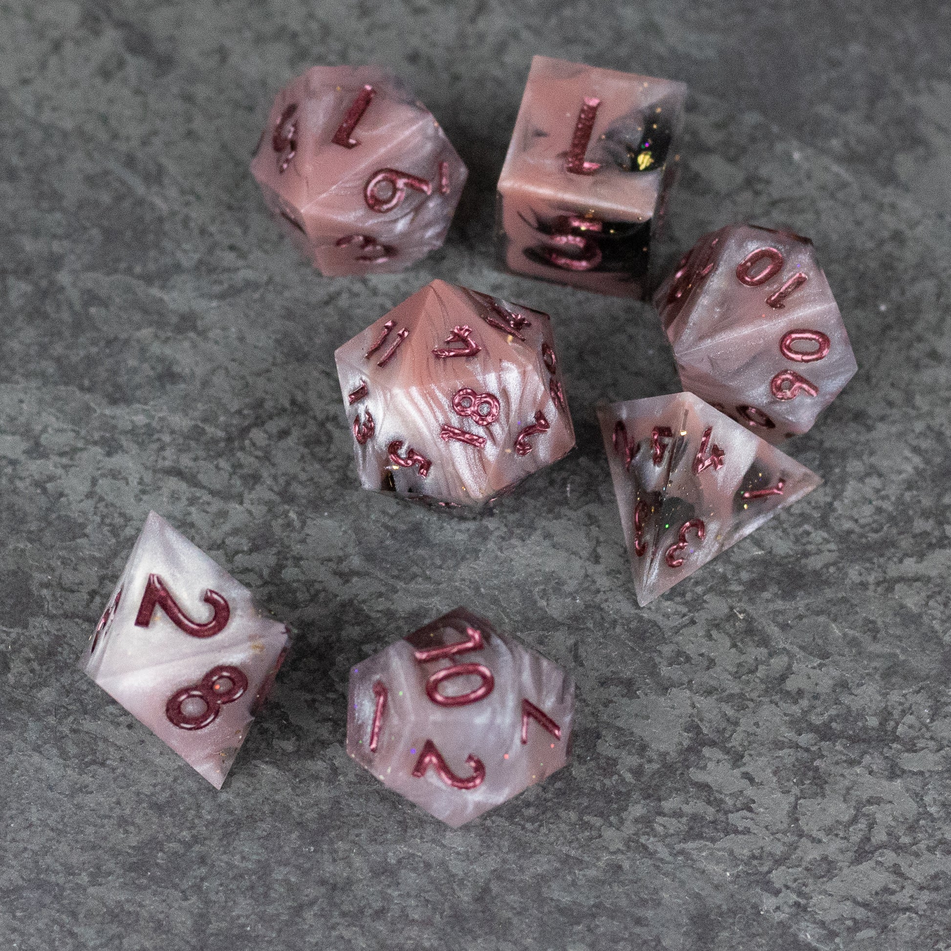 Rosestone Dice Set - Seven red and pink marbled sharp-edged polyhedral dice with white and black numbers on a gray textured surface.